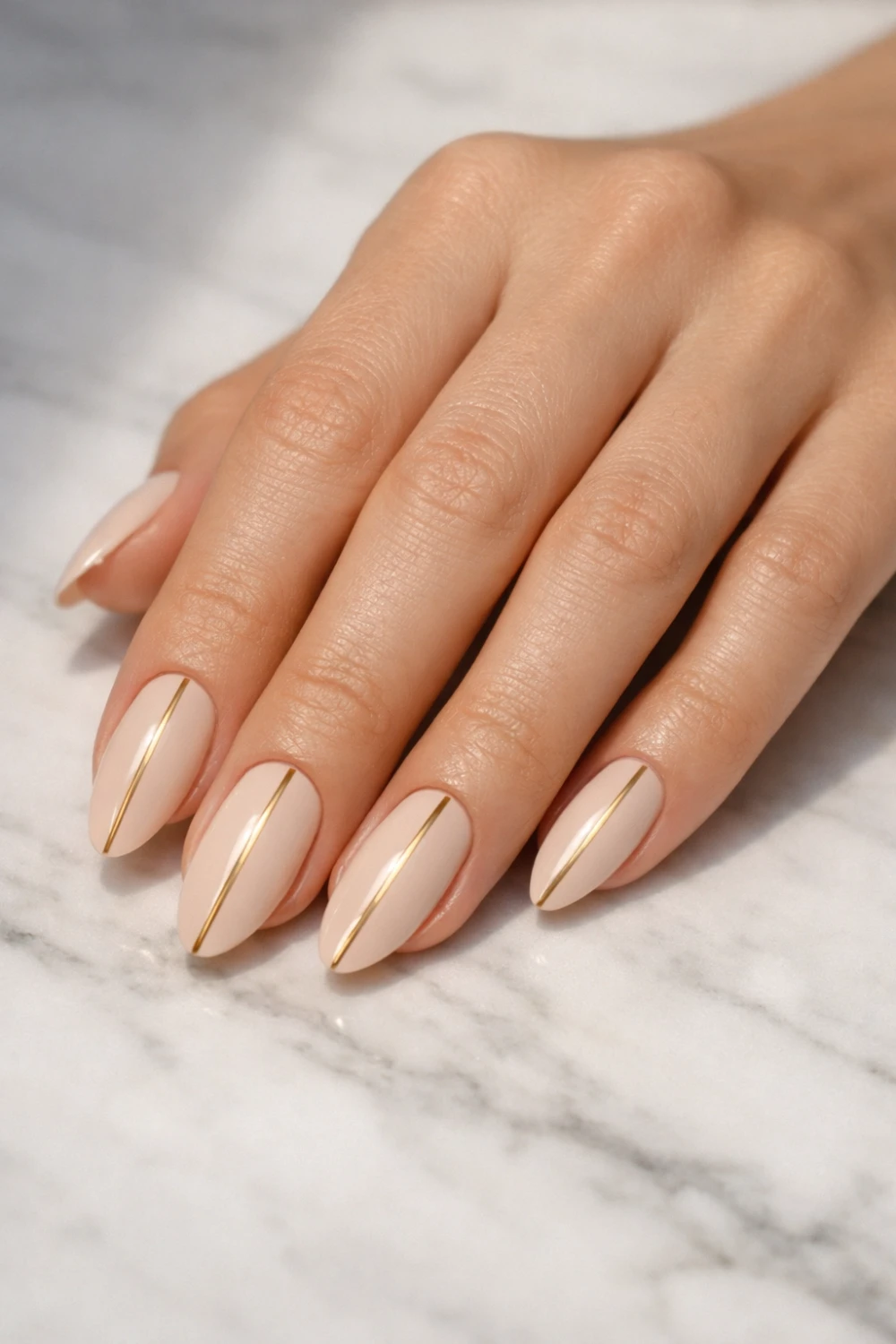 Minimal nude almond nails with thin gold vertical lines on marble background showcasing modern old money manicure style