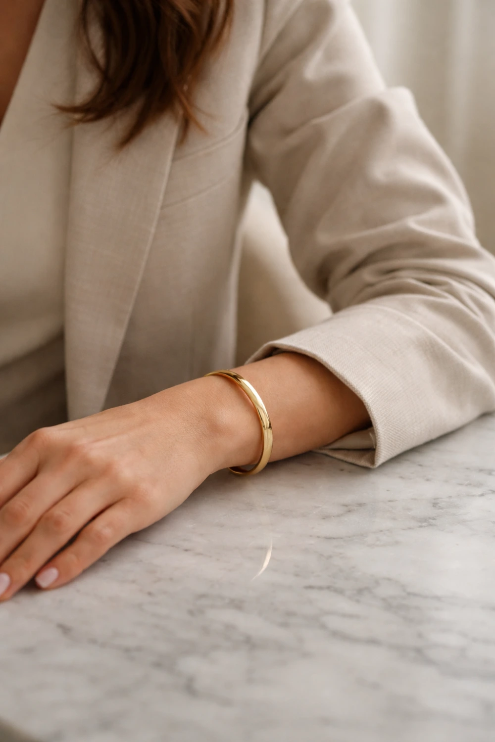 Woman wearing a polished gold bangle bracelet with a neutral blazer sleeve resting on a marble table, highlighting a timeless old money jewelry look with elegant, minimal, and quietly luxurious styling.