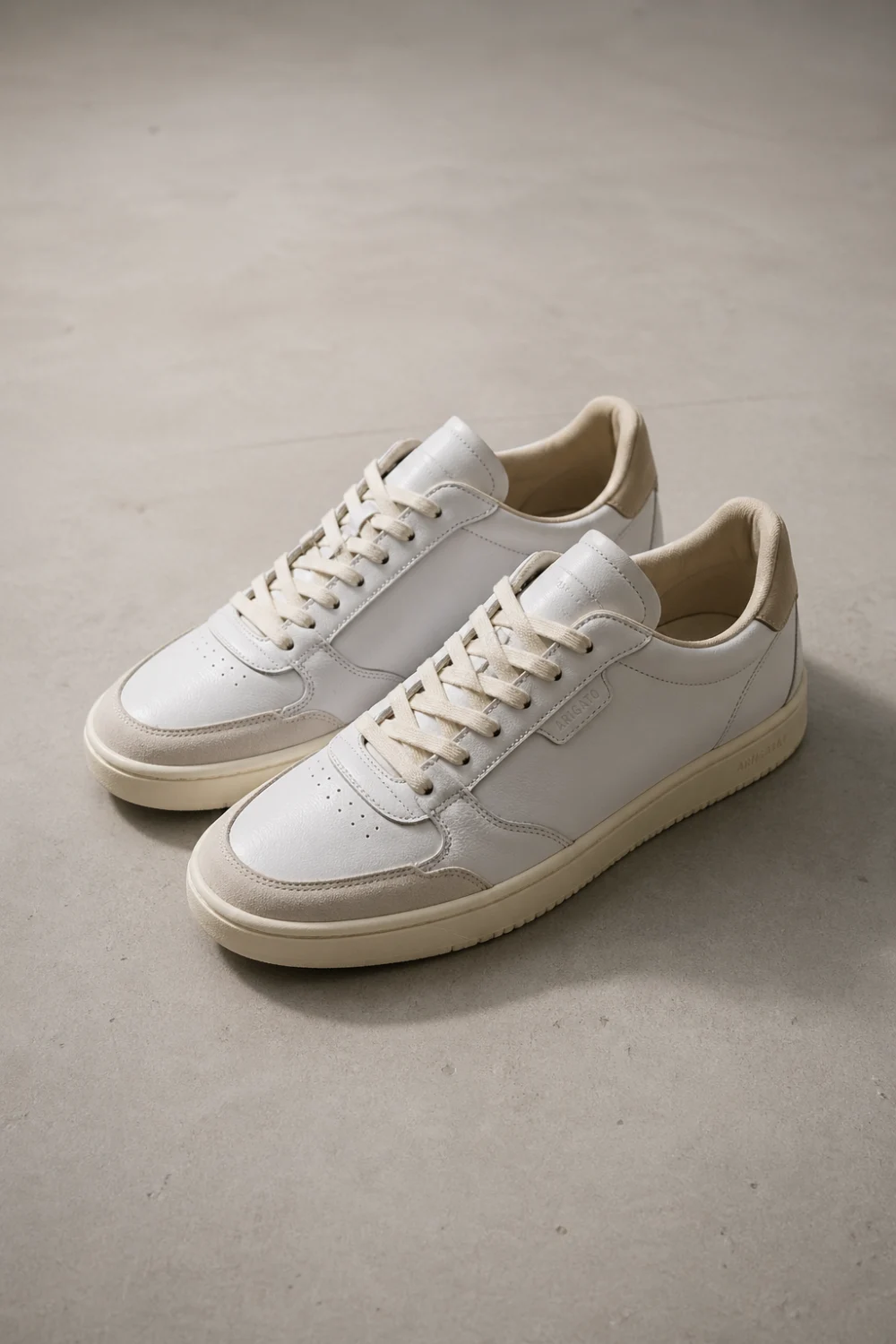 Clean white slim minimal sneakers placed on a neutral beige surface, modern low profile design with lace up detail, soft natural lighting, minimalist aesthetic, showcasing everyday casual footwear trend for effortless street style looks