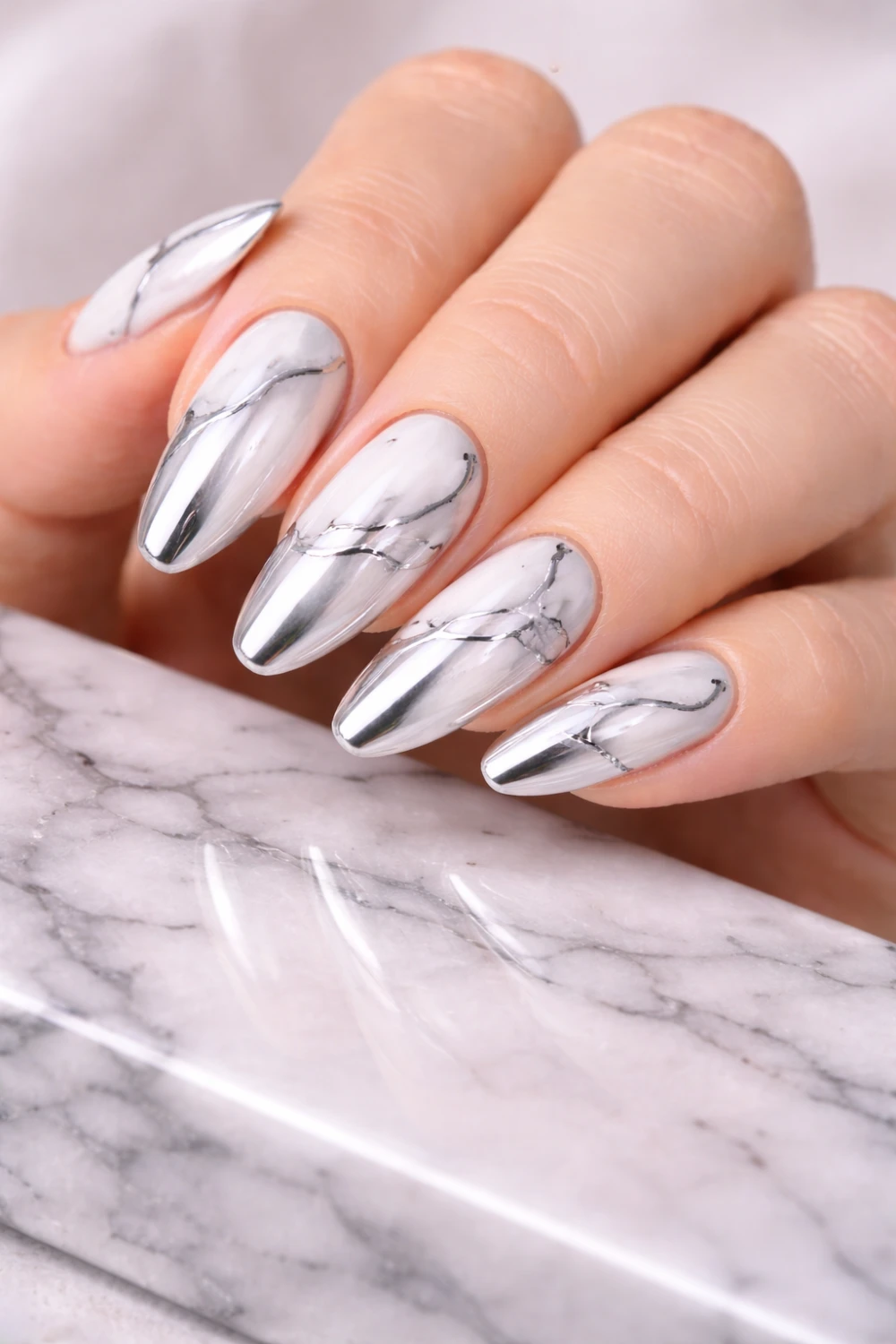 Chrome marble nails with white marble pattern and metallic silver accents on almond manicure, glossy marble chrome nail design photographed on marble surface