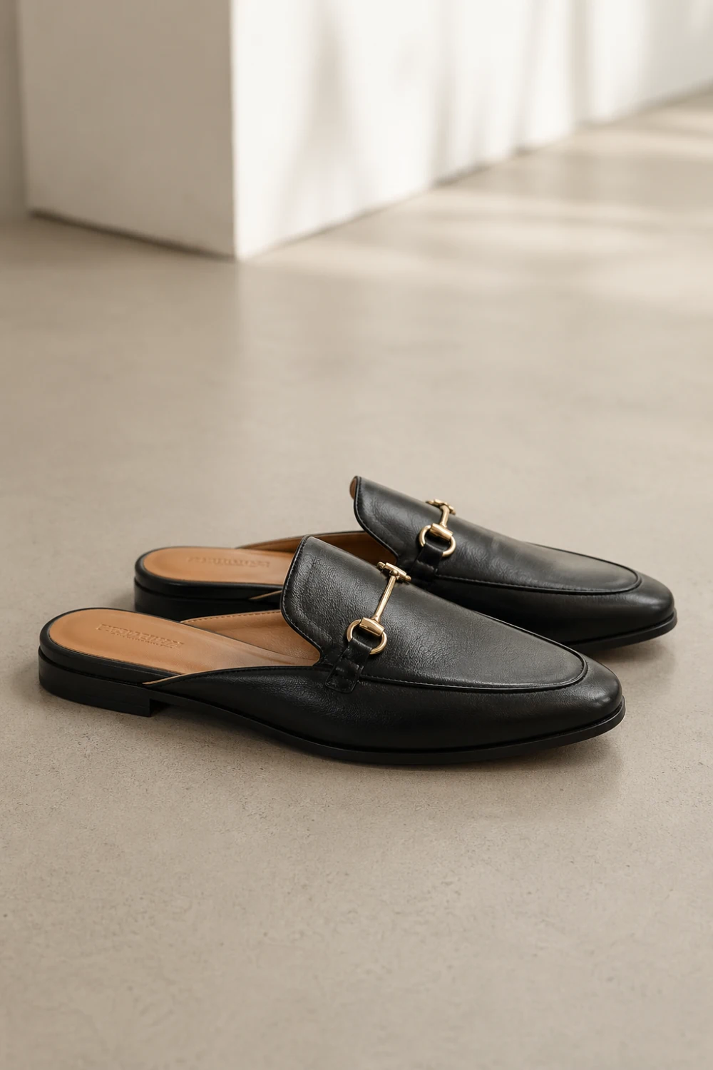 Black backless loafers with gold detail placed on neutral floor, slip on mule style shoes, minimal modern footwear, clean and polished look perfect for casual and smart outfits
