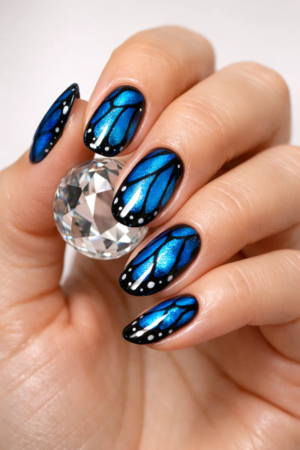 Elegant female hand holding crystal showing blue morpho butterfly wing nail art with metallic blue shimmer and black wing patterns, glossy gel manicure butterfly nails inspiration