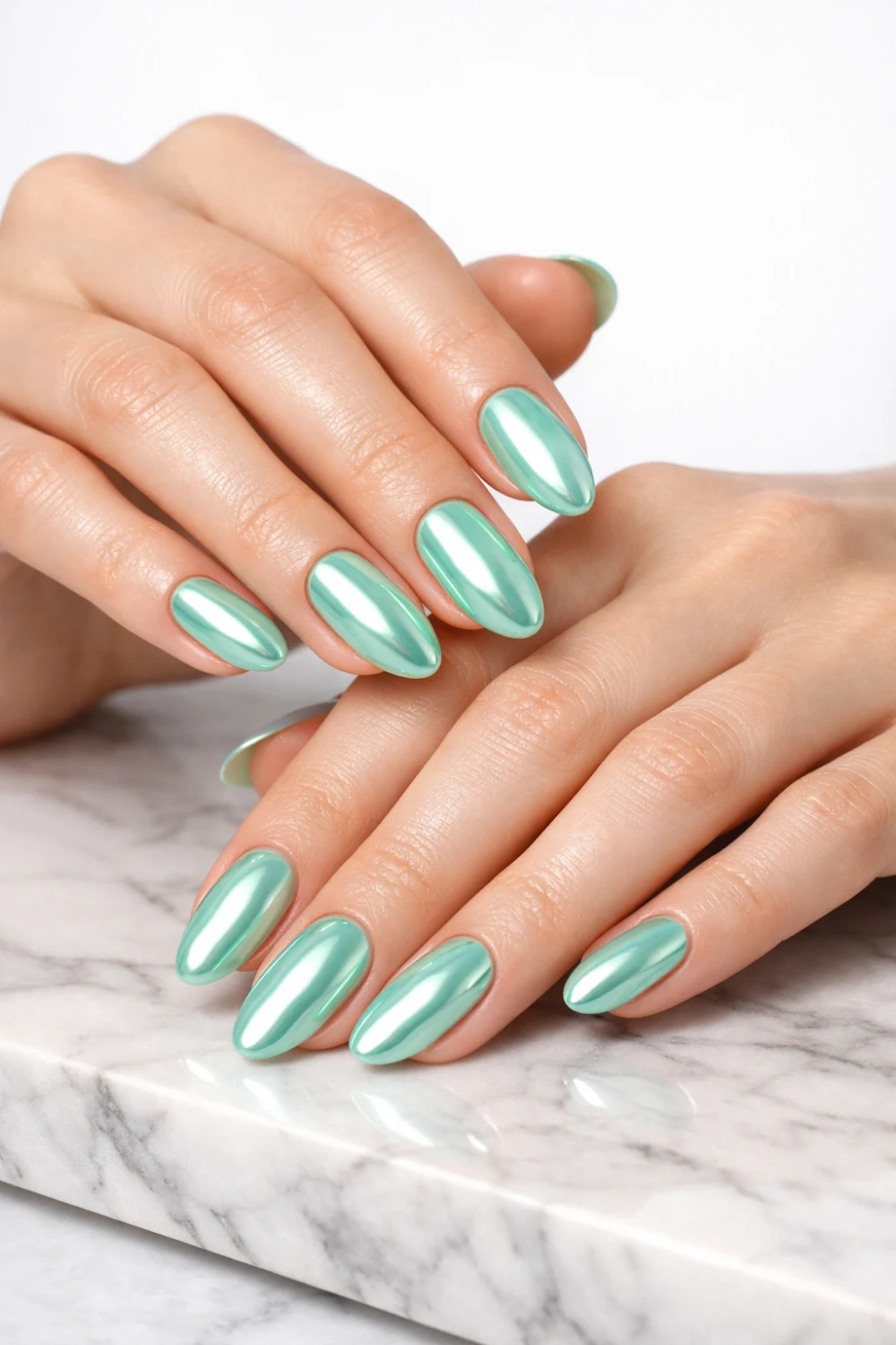 Elegant feminine hands with oval-shaped mint green chrome nails, reflective glossy finish, smooth skin, minimal marble surface background, modern pastel nail design