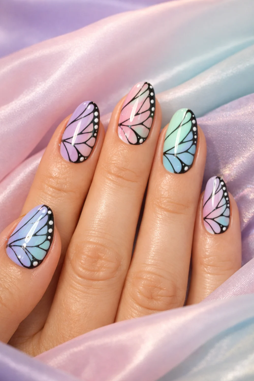 Full female hand over pastel fabric showing pastel butterfly wing nail art in lavender pink mint and blue colors, glossy gel manicure dreamy butterfly nail design inspiration