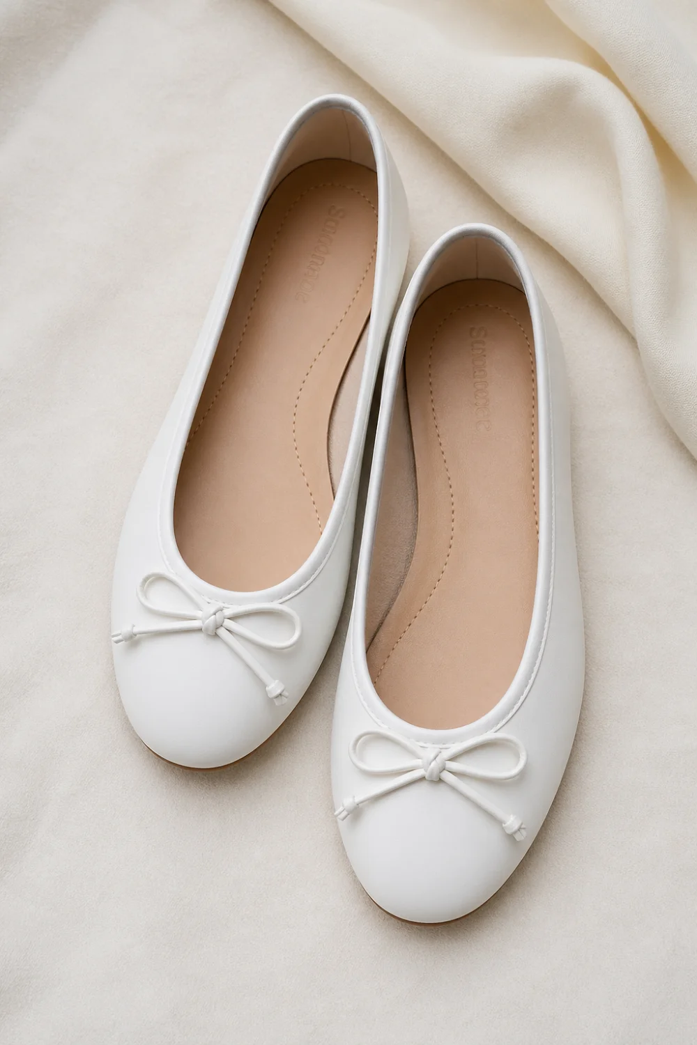 White flat ballet shoes with soft rounded toe and bow detail placed on neutral fabric background, clean minimalist footwear style, lightweight everyday shoes perfect for simple chic outfits and modern wardrobe essentials