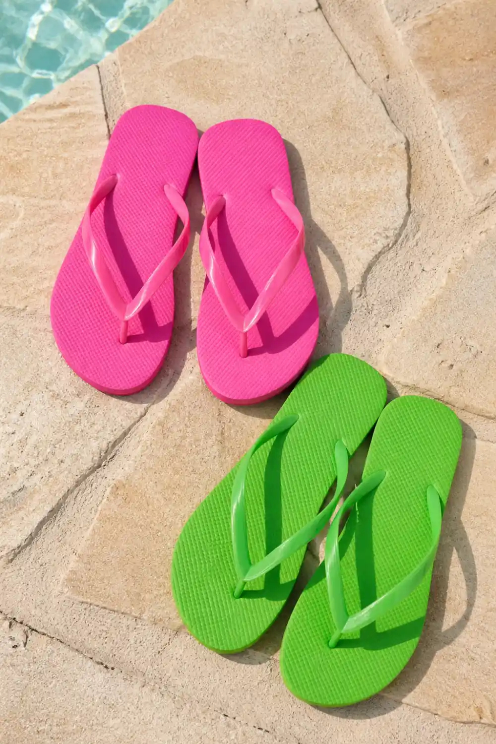 Bright pink and green colorful flip flops placed on sunlit stone surface near pool, vibrant summer footwear, casual beach style sandals, bold trendy look perfect for relaxed outfits and vacation vibes