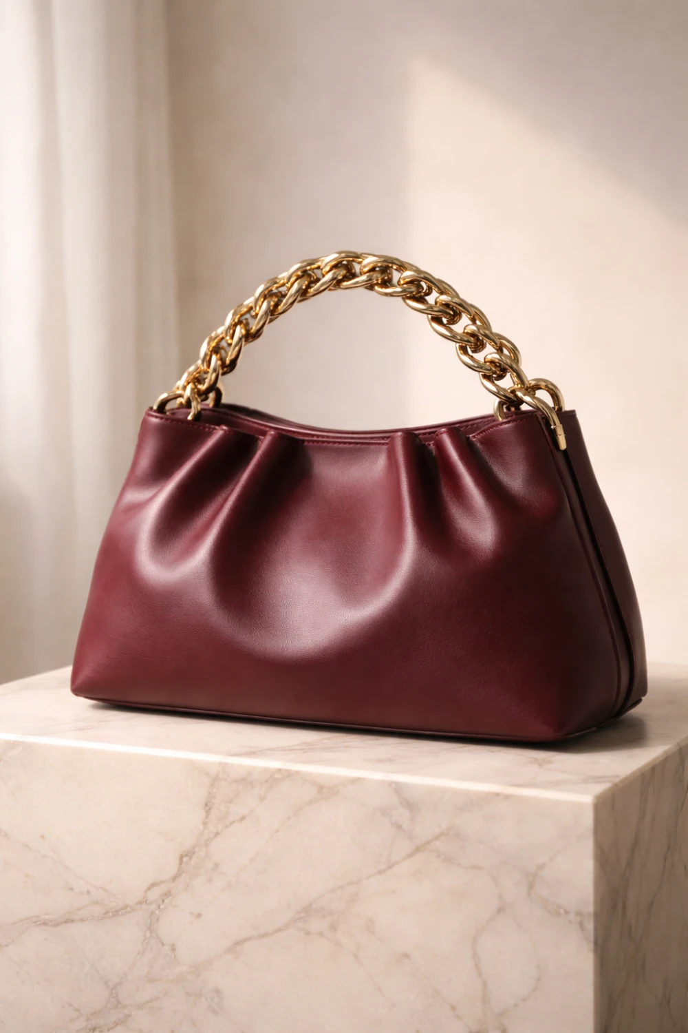 Burgundy leather handbag with chunky gold chain handle placed on marble display, luxury chain strap handbag trend for women fashion 2026.