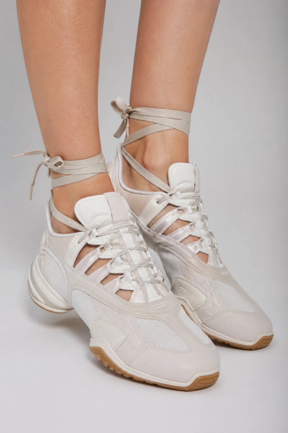 White ballet sneakers with lace up straps wrapped around ankles, hybrid sporty and feminine design, modern fashion footwear, soft neutral tone sneakers styled for trendy streetwear looks