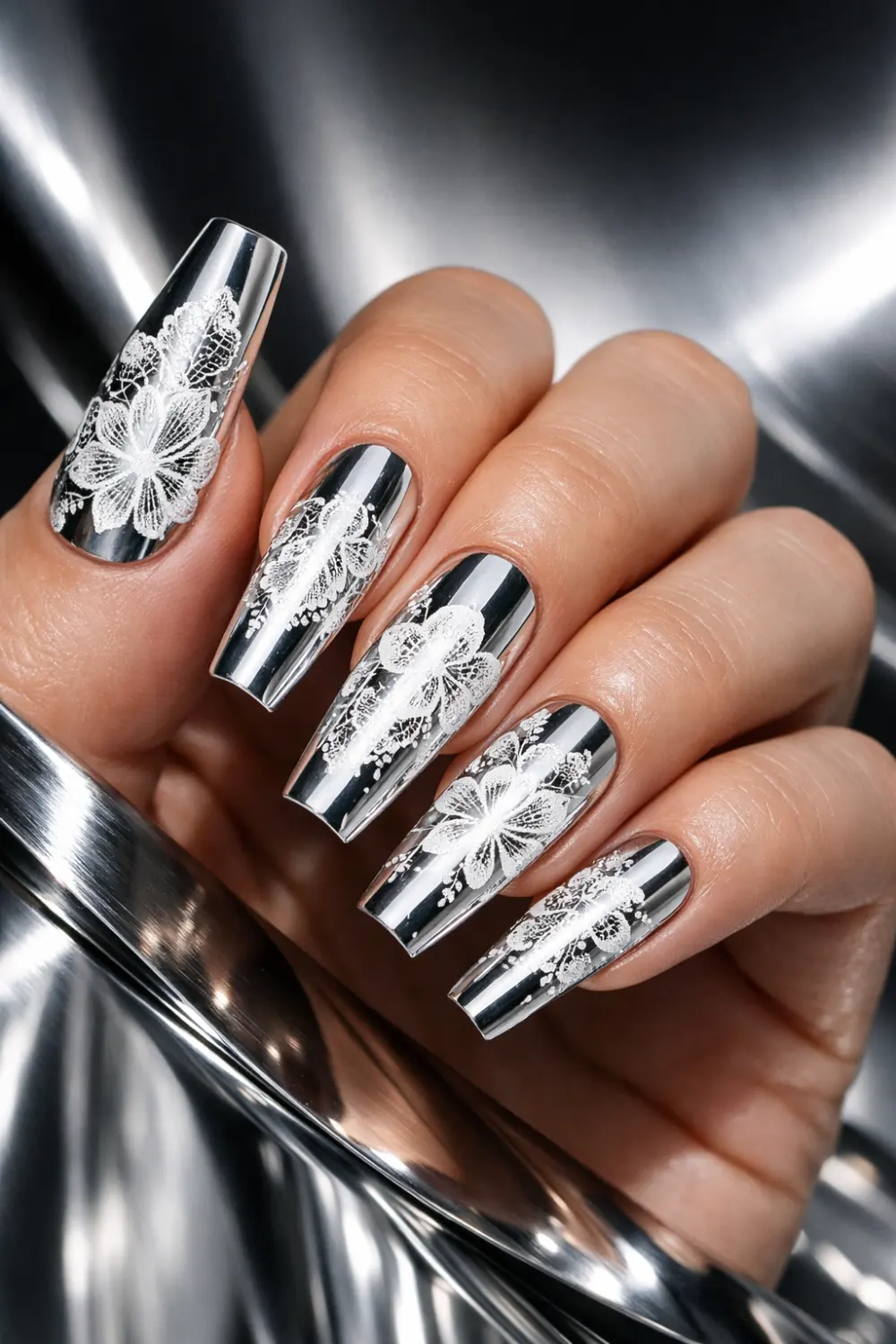 Mirror chrome manicure with intricate white lace floral nail art over metallic silver nails, glamorous coffin shaped nails with glossy reflective finish creating a luxurious chrome lace nail design perfect for statement manicure inspiration