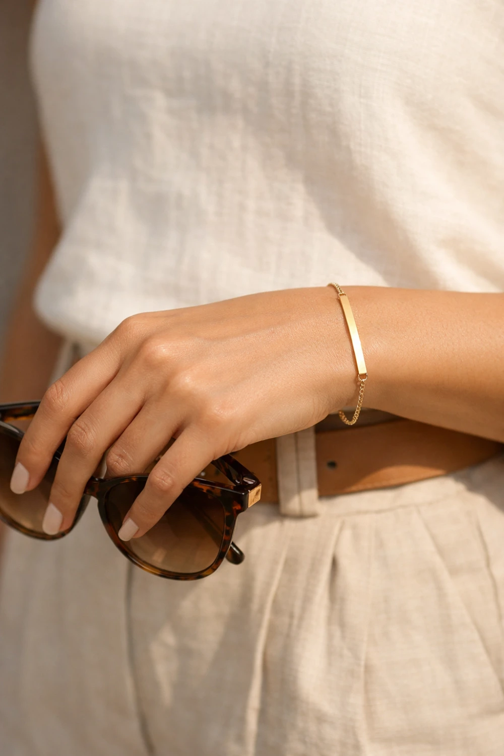 Minimal gold bar bracelet worn on a woman's wrist while holding sunglasses, showing a modern yet elegant old money jewelry aesthetic with clean minimalist styling.