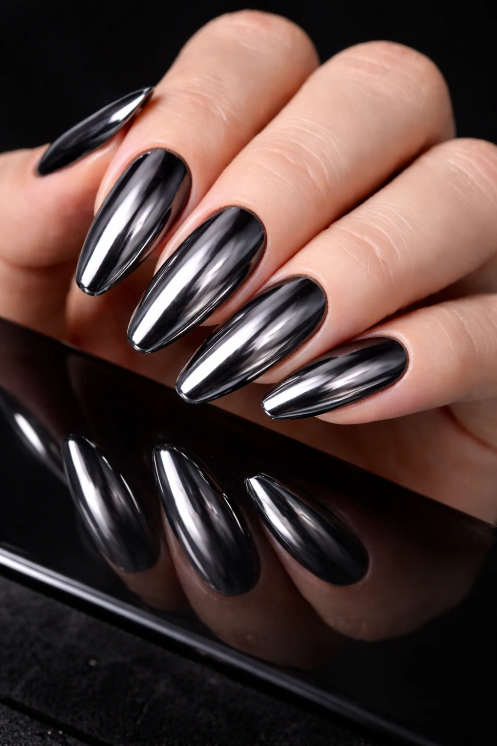 Black chrome mirror nails with deep metallic shine on almond shaped manicure, glossy reflective dark chrome nail design photographed on black background, edgy chrome nail inspiration