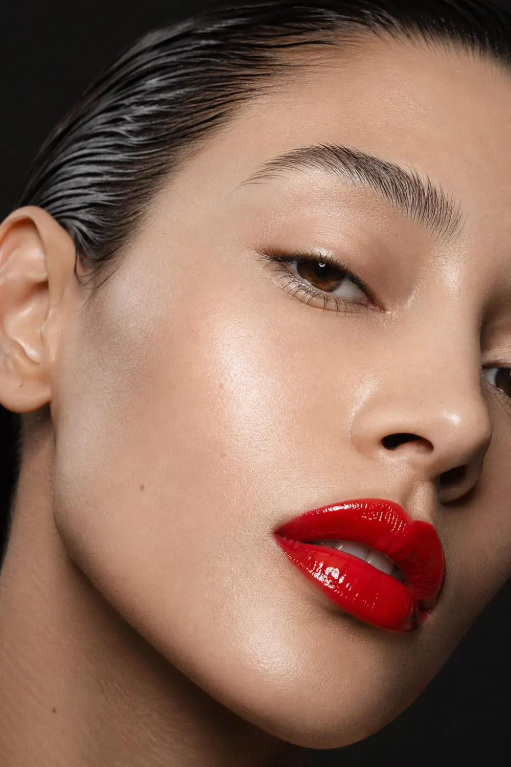 Close up beauty portrait with glossy high shine red lipstick, slicked back wet hair, radiant glowing skin, reflective lip texture, dark background, luxury editorial makeup style