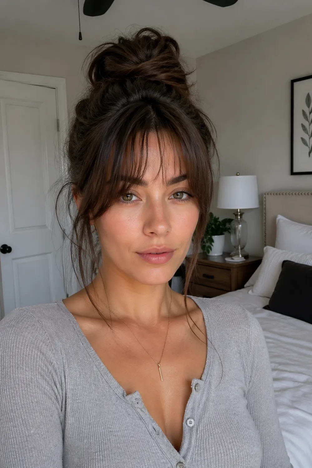 messy high bun hairstyle with curtain bangs loose strands soft face framing head to chest portrait in bedroom interior with bed lamp and cozy natural lighting