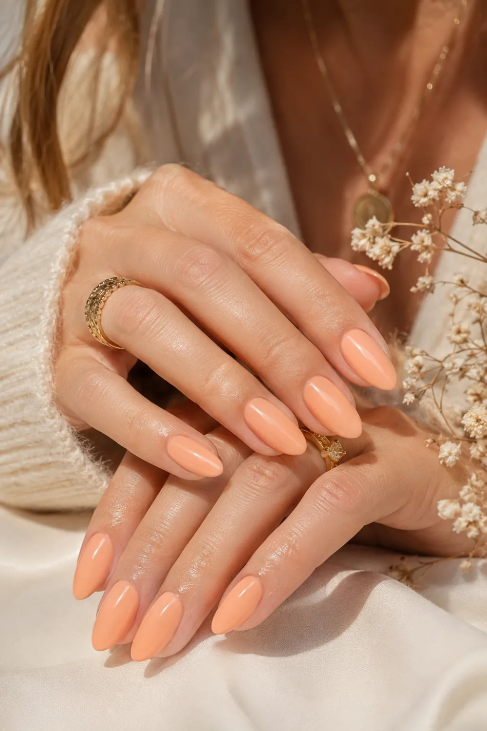 Elegant feminine hands with glossy soft peach almond nails, gold rings, cozy cream sweater styling, and classy clean girl summer manicure aesthetic