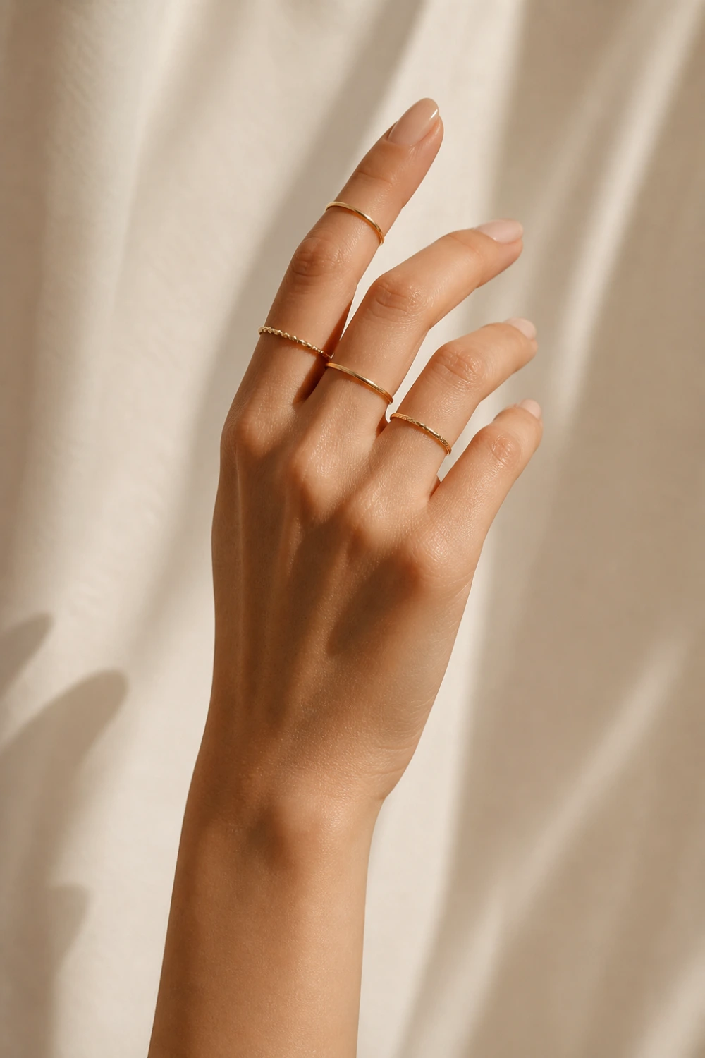 close-up female hand wearing thin minimal gold knuckle rings stacked across upper fingers, clean nude manicure, soft neutral beige background, luxury minimalist jewelry aesthetic, soft natural lighting, elegant modern midi rings style