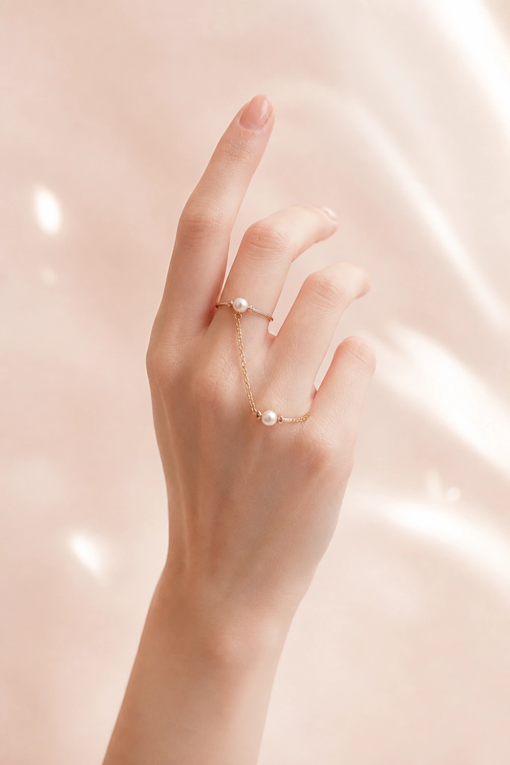 female hand wearing pearl knuckle rings with delicate gold chain detail, soft pastel background, feminine jewelry aesthetic, minimal manicure, elegant midi rings, luxury soft style