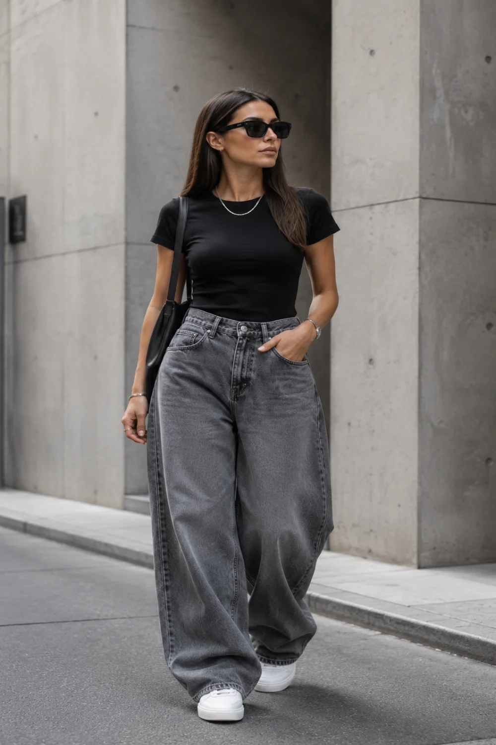 Minimalist woman wearing a fitted black t-shirt with oversized gray baggy denim jeans, white sneakers, slim sunglasses, and a black shoulder bag against a modern concrete city background.