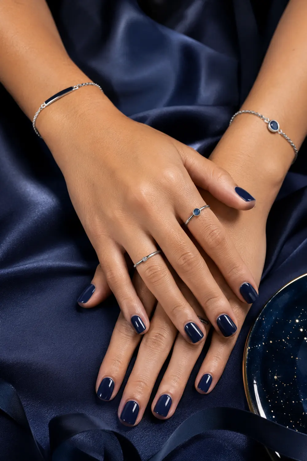 Glossy navy blue short nails with elegant satin background, silver jewelry accents, luxury beauty styling, and sophisticated feminine manicure aesthetic.