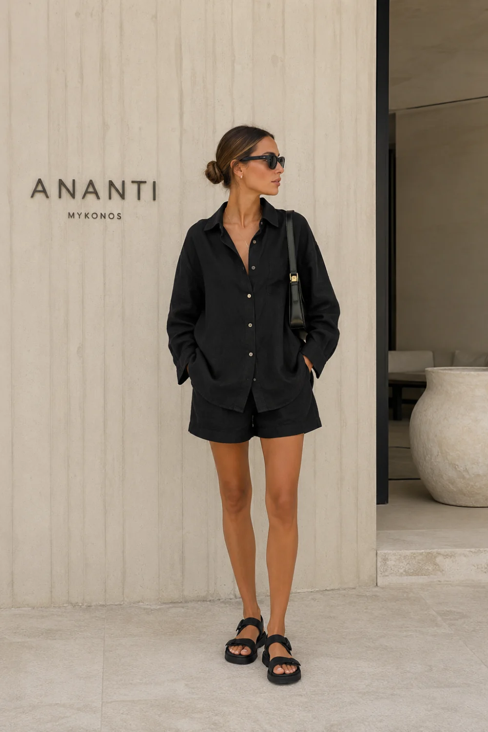 Woman wearing a black oversized linen shirt and matching shorts co-ord set with chunky sandals and sunglasses in a luxury minimalist setting