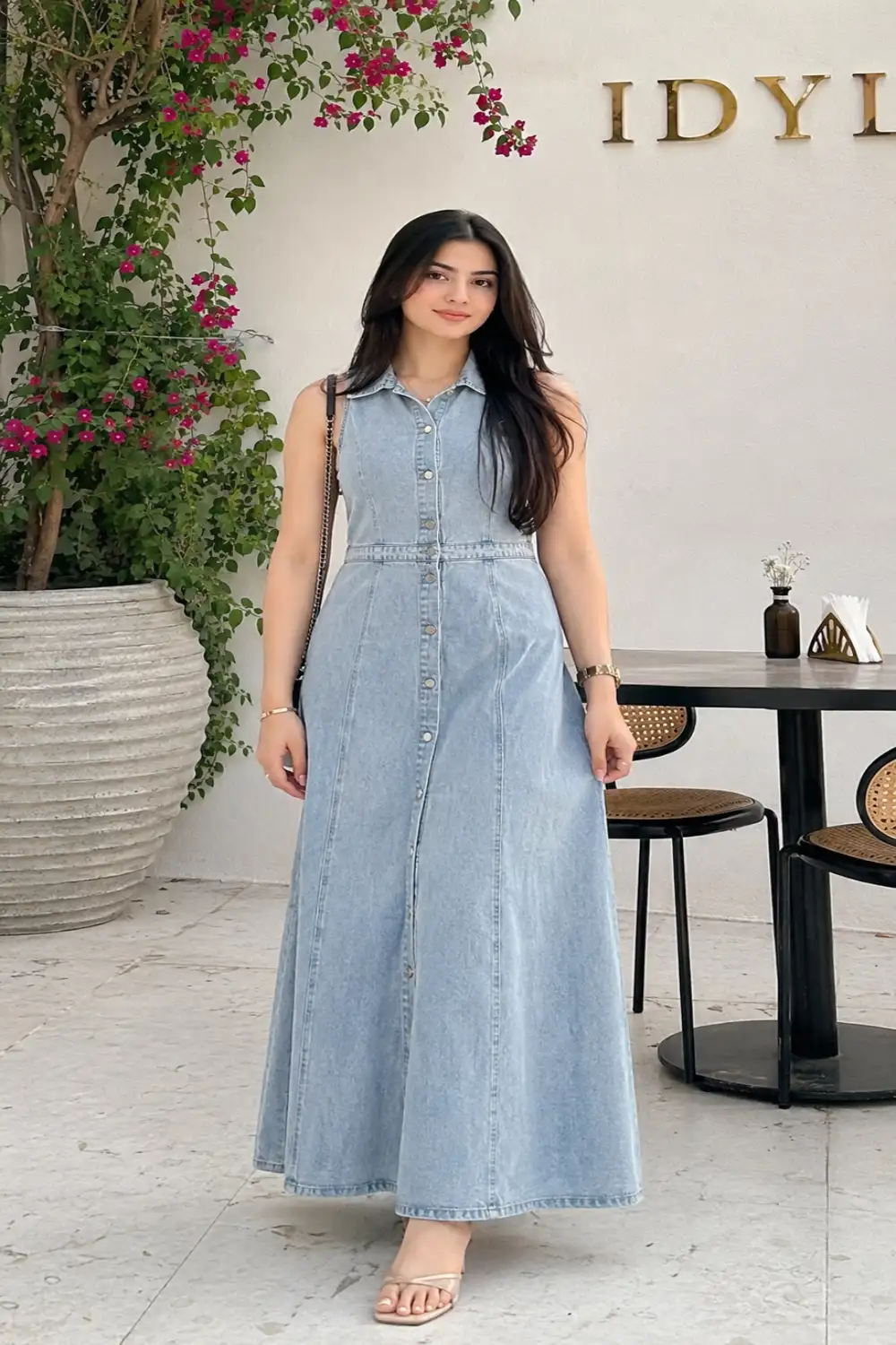 Woman wearing a sleeveless light wash denim maxi dress with flat sandals and a shoulder bag in a beautiful outdoor café setting.