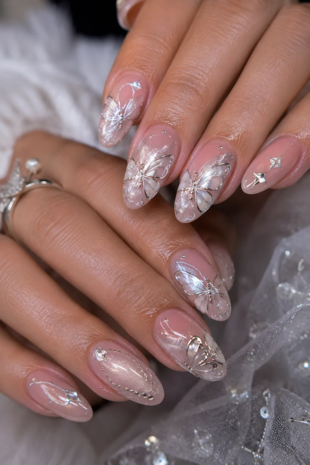 Silver butterfly glam almond nails with glossy nude pink gel polish, sparkling rhinestone details, elegant butterfly wing nail art, and luxury manicure design.