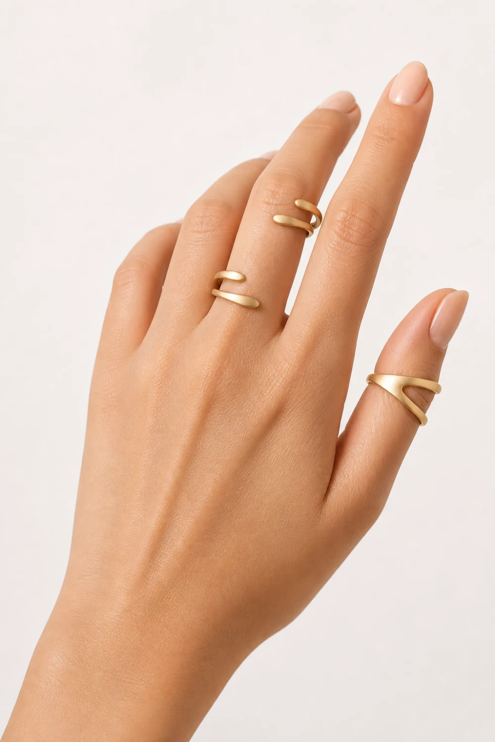 female hand wearing open adjustable knuckle rings with smooth wrap design in matte gold, minimalist midi rings, clean white background, modern jewelry aesthetic, soft natural lighting