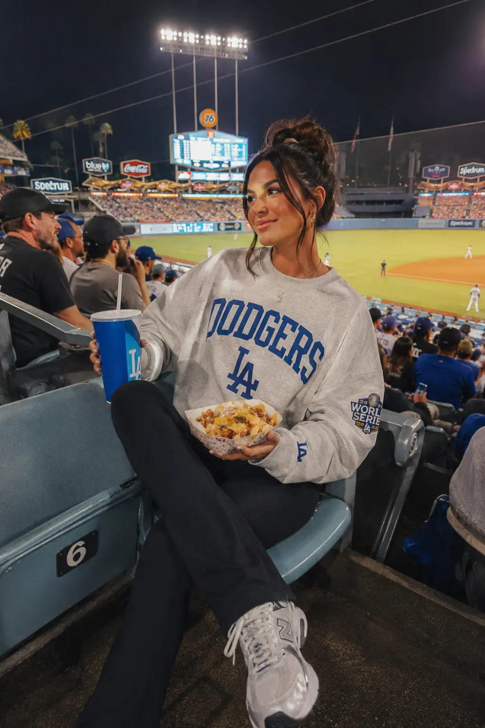 Woman wearing oversized Dodgers sweatshirt with black flare leggings and chunky sneakers sitting in stadium seats during night baseball game.