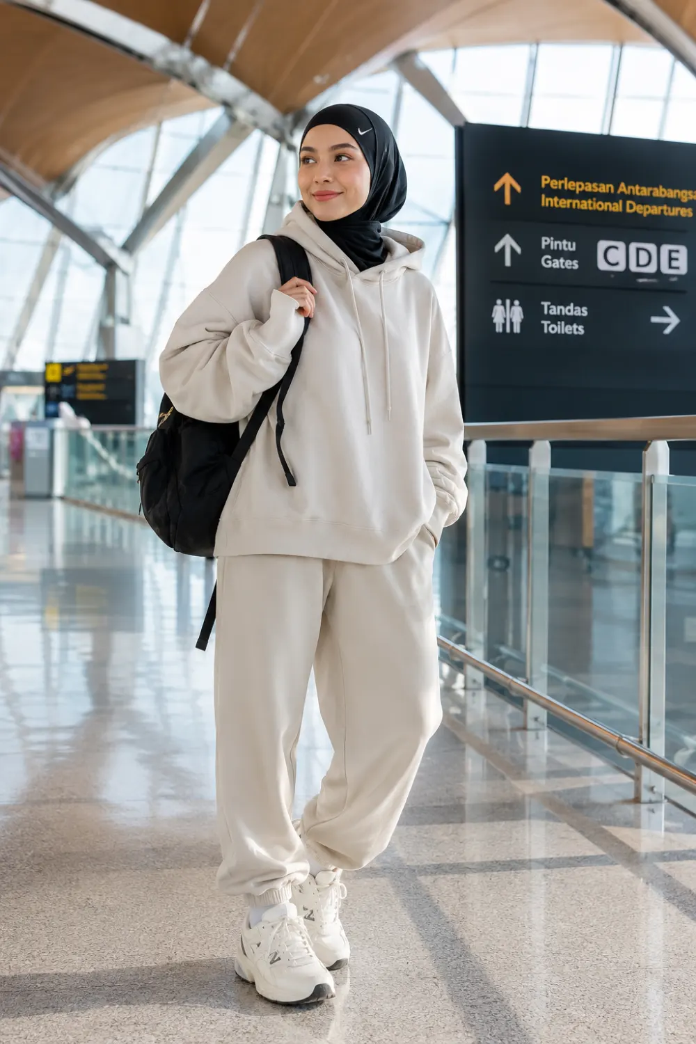 Cute sporty hijabi woman wearing oversized neutral joggers, matching hoodie, chunky sneakers, black sports hijab, and a backpack while walking through a modern airport terminal. The modest athleisure outfit features cozy travel fashion, relaxed neutral tones, and trendy sporty styling perfect for everyday comfort.