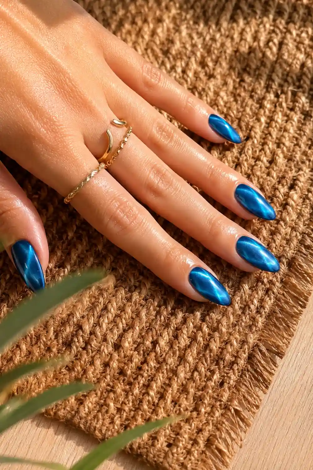 Elegant ocean blue cat eye nails with a glossy magnetic shimmer effect on almond-shaped nails, photographed on a feminine hand resting on a brown woven rug over a light wood table with delicate gold rings and warm summer sunlight.