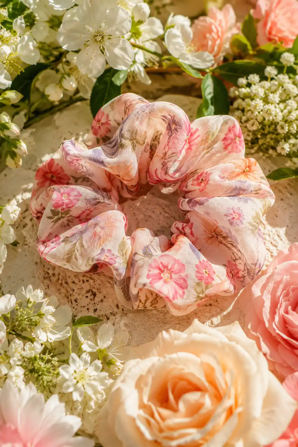 Romantic floral chiffon scrunchie featuring soft pink floral prints displayed among blooming roses, garden flowers, and warm natural sunlight. The lightweight chiffon fabric creates an airy feminine look, making it a beautiful accessory for spring hairstyles, summer fashion, vacation outfits, and floral-inspired styling.