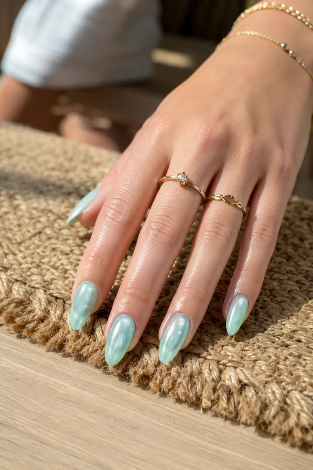 Mint green cat eye nails with soft magnetic shimmer on almond-shaped nails, displayed on a feminine hand with gold rings resting on a woven rug and wooden table.