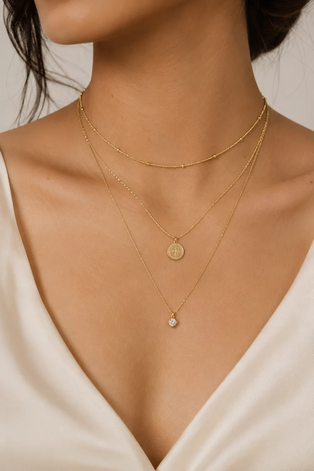 Woman wearing delicate layered gold necklaces with a coin pendant and crystal charm styled with a soft ivory satin top for an elegant jewelry look.