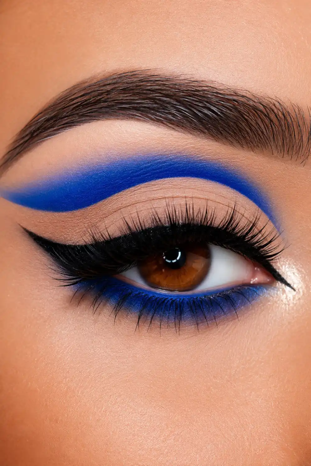 Electric blue cut crease eye makeup featuring vivid cobalt blue eyeshadow, sharp crease definition, dramatic black winged eyeliner, voluminous lashes, luminous inner corner highlight, and glamorous beauty makeup for parties and special occasions.