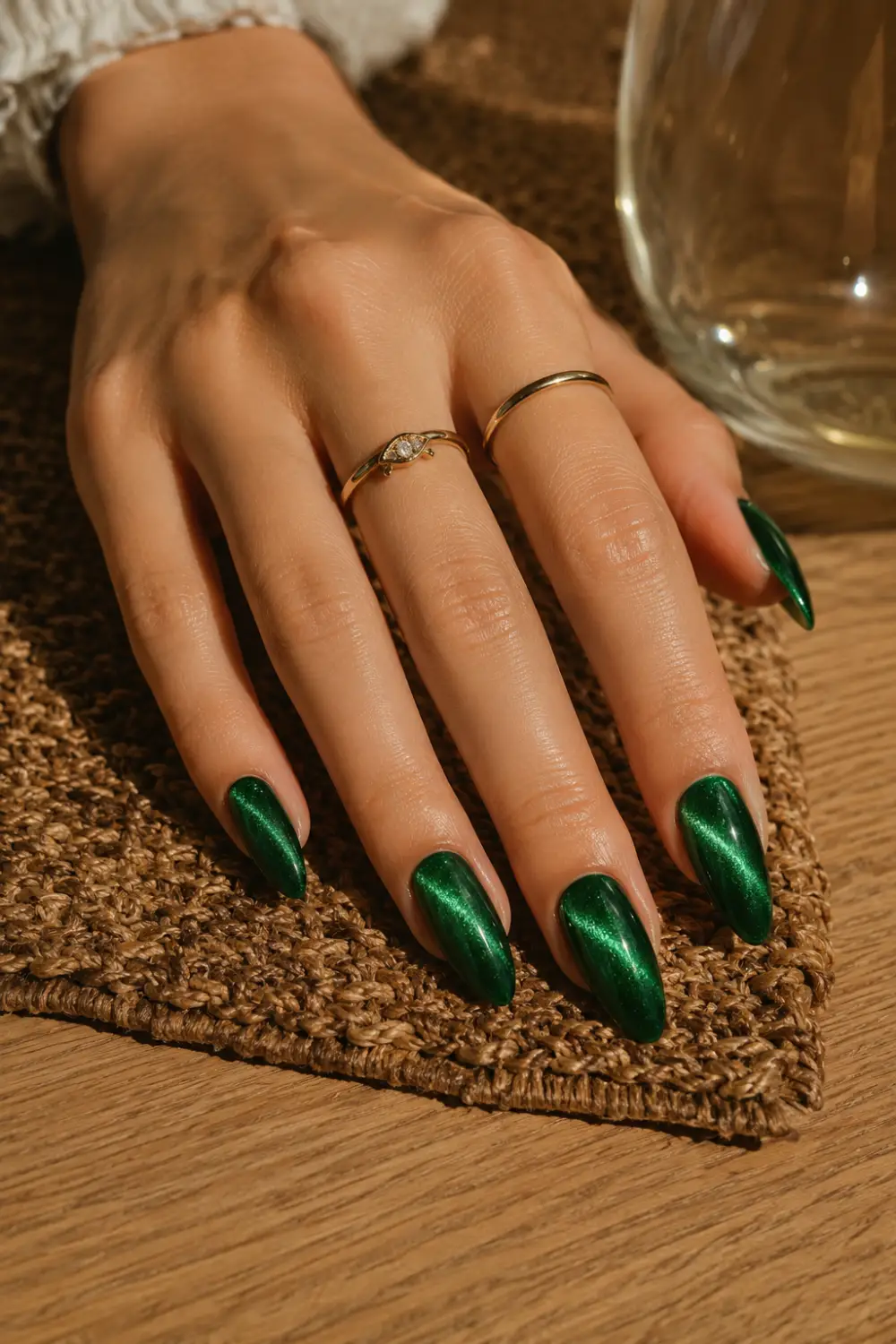 Luxurious emerald green cat eye nails with rich magnetic shimmer on almond-shaped nails, photographed on a feminine hand resting on a textured brown woven rug with delicate gold jewelry.