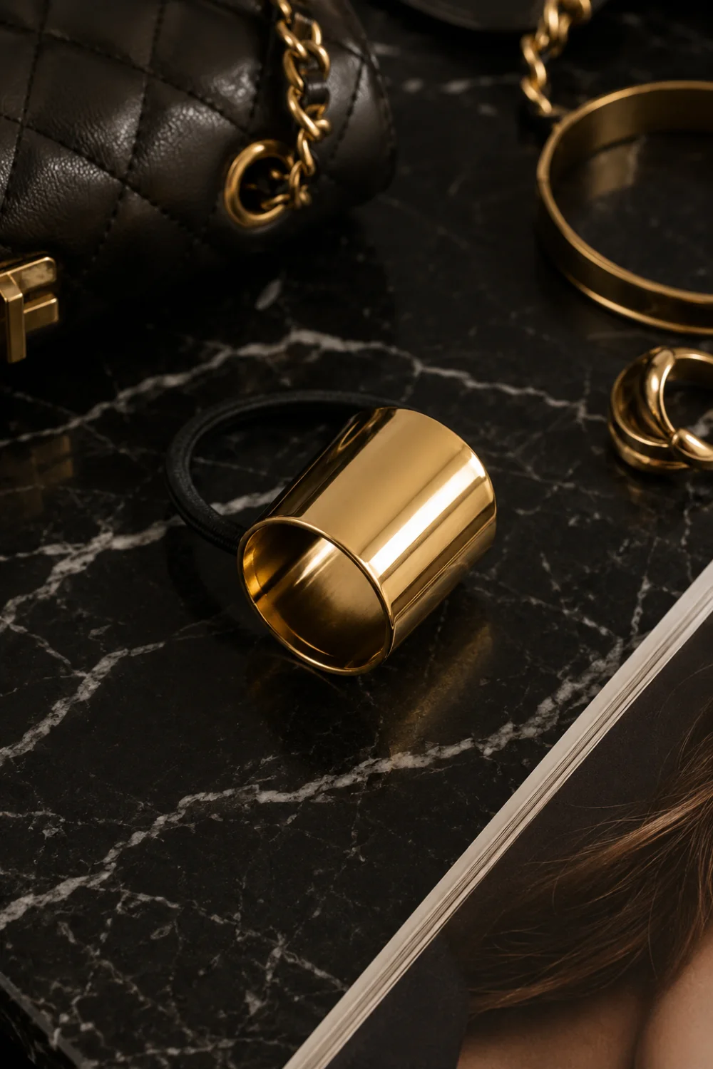 Modern gold metal hair cuff ponytail holder with a polished reflective finish displayed on black marble alongside designer-inspired accessories, gold jewelry, and luxury fashion details. The sleek metallic design creates a sophisticated statement hairstyle while adding a contemporary high-end aesthetic to any ponytail.