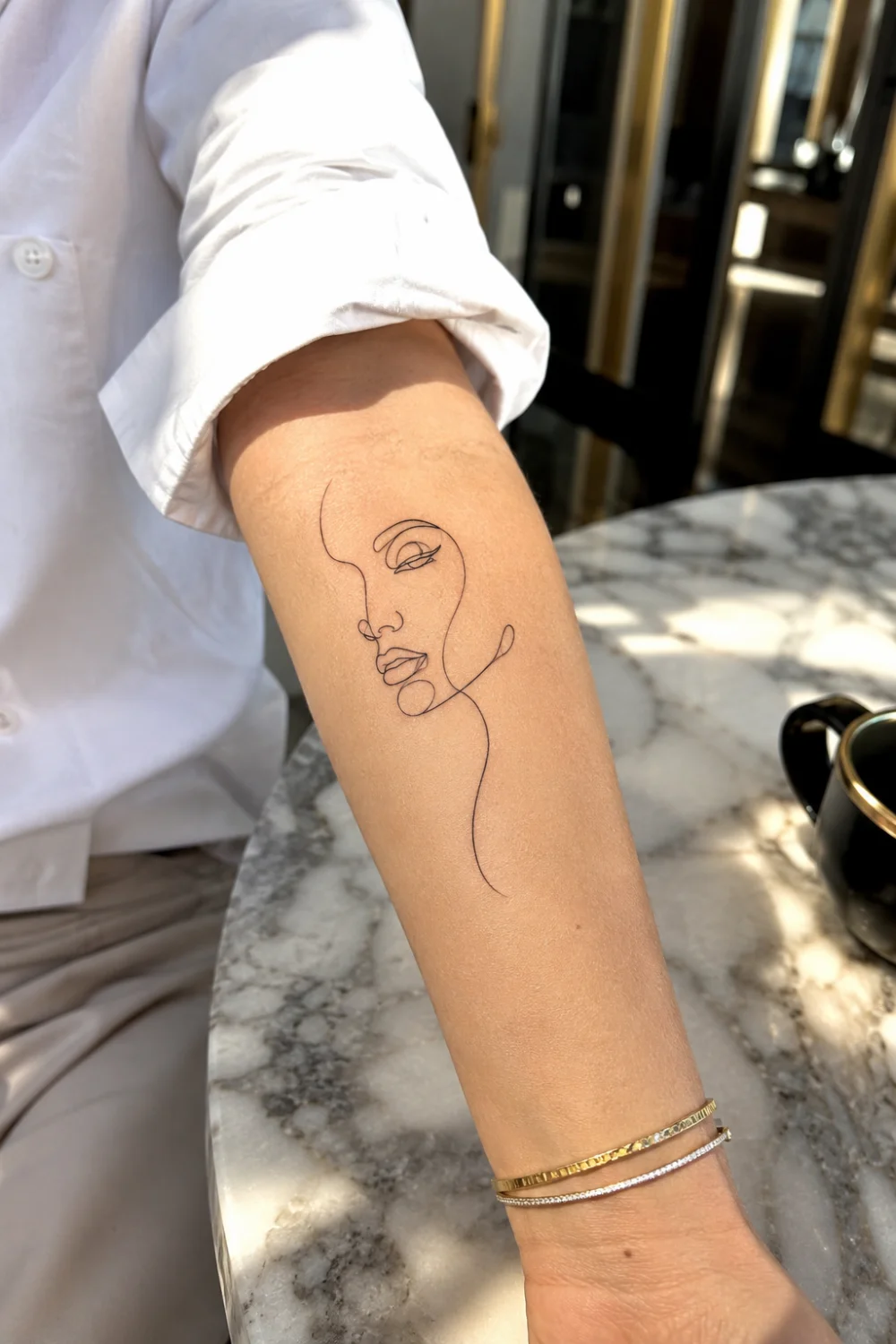 Female face line art tattoo on a woman's forearm featuring continuous line drawing artwork, abstract feminine tattoo design, minimalist black ink illustration, modern aesthetic tattoo inspiration, and artistic body art.