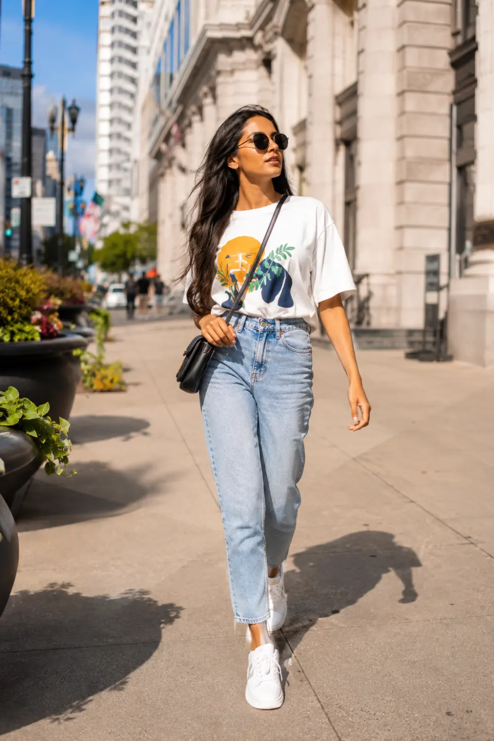Fashionable Indian woman wearing a graphic t-shirt tucked into light wash mom jeans with white sneakers and sunglasses, walking confidently through a modern city street, trendy casual summer outfit inspiration.