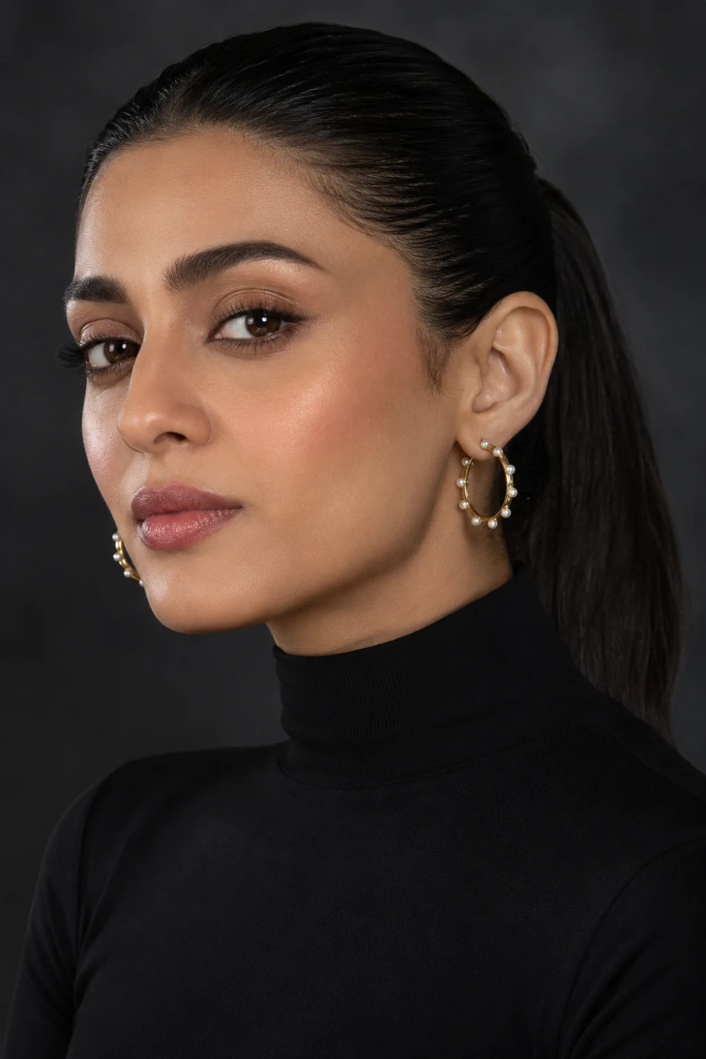 Close up portrait of a woman wearing pearl hoop earrings with delicate pearl accents, sleek hairstyle, black turtleneck outfit, and modern luxury styling.