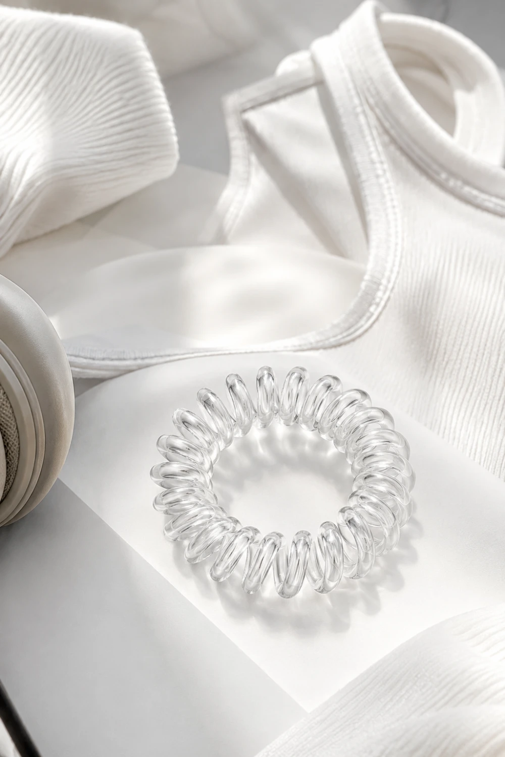 Transparent spiral hair tie with a crystal-clear coiled design displayed beside activewear, headphones, and minimalist fashion accessories. The telephone-cord shape provides a secure hold while reducing hair creases, making it a stylish and practical accessory for everyday ponytails, gym hairstyles, and modern casual looks.