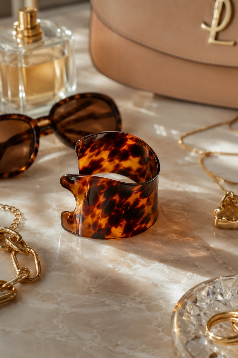 Luxury tortoise shell ponytail cuff featuring rich amber and brown marbled patterns displayed on a designer-inspired vanity with sunglasses, perfume, gold jewelry, and elegant accessories. The glossy finish and timeless pattern create a sophisticated ponytail accessory that complements both casual and formal fashion.