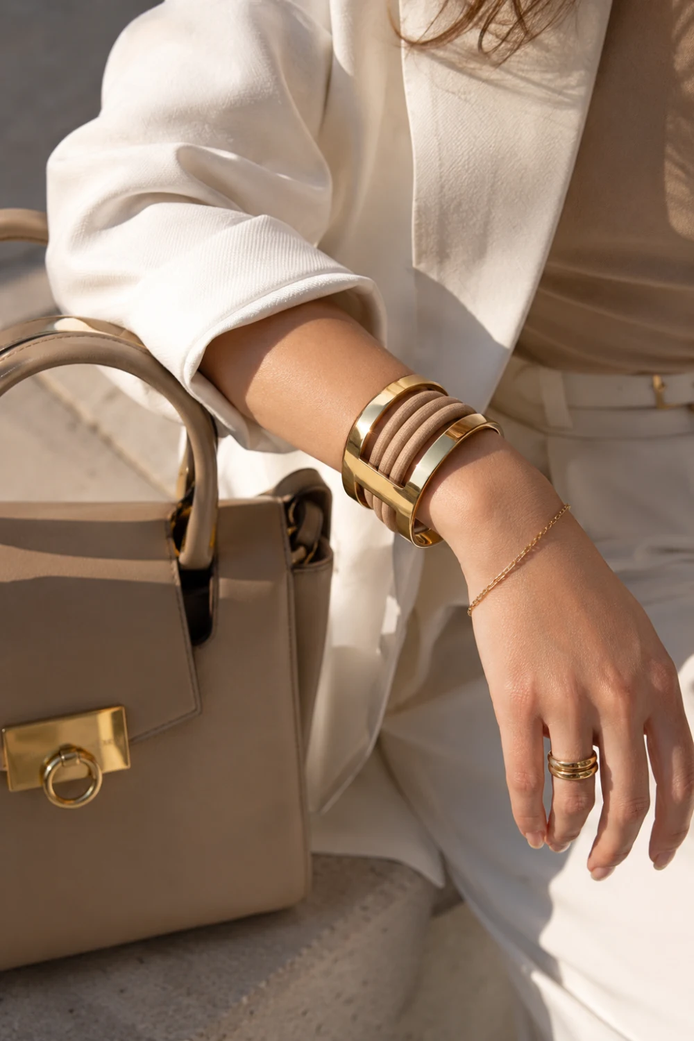 Elegant gold hair tie bracelet holder worn on a fashionable wrist alongside luxury jewelry, a designer handbag, and neutral-toned clothing. The sleek metallic design doubles as a bracelet and hair accessory holder, offering a stylish solution for carrying hair ties while maintaining a polished appearance.